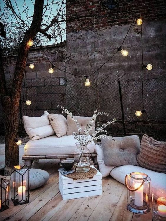 a Scandinavian winter terrace with a loveseat, lots of cushions and pillows, candle lanterns, a bulb garland and some blooms