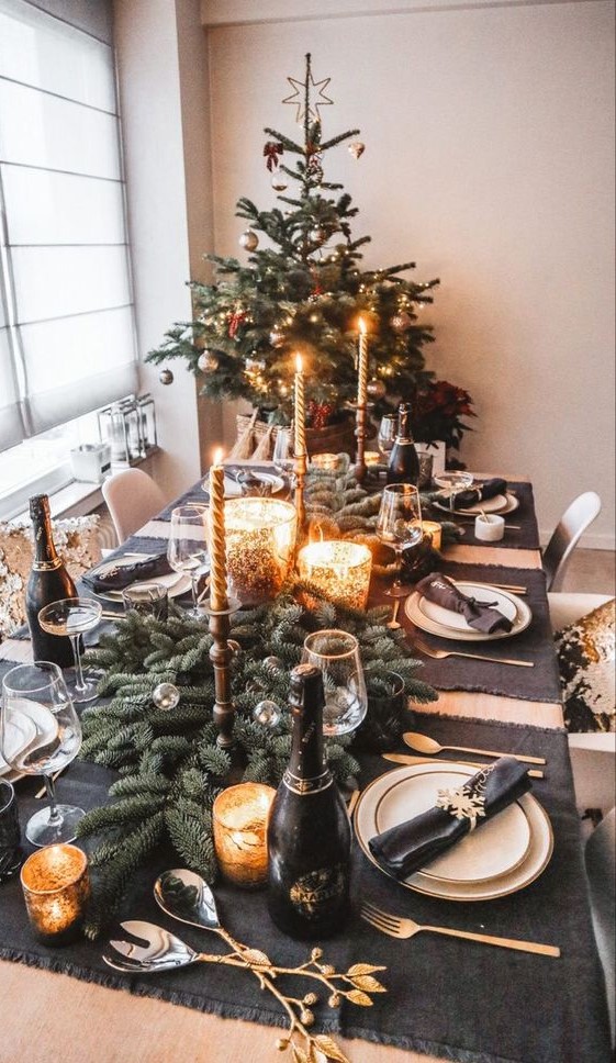 a black and gold party table with dark linens, white porcelain, gold cutlery, candleholders and evergreens
