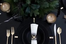 a black table with black plates and cahrgers, lush evergreens and gold cutlery for a moody tablescape