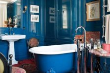 a bold bathroom with shiny paneled blue walls, a bold blue and red printed rug, a blue clawfoot bathtub and vintage furniture