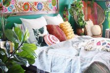 a bold boho bedroom with emerald walls, a bright artwork, a bed with lots of mismatching pillows, potted plants and a macrame lamp hanging
