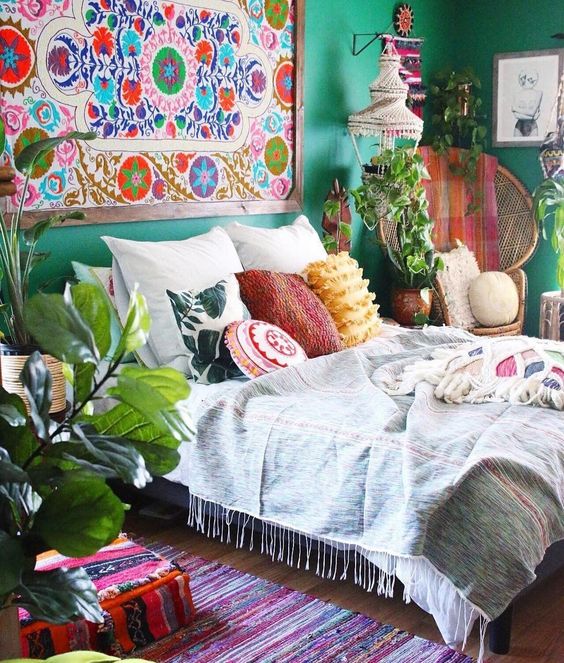 a bold boho bedroom with emerald walls, a bright artwork, a bed with lots of mismatching pillows, potted plants and a macrame lamp hanging