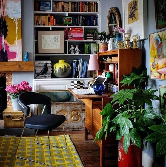 a bold maximalist home office with a blue wall, a fireplace, a vintage bureau desk, colorful artworks and potted plants