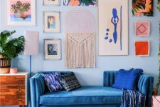 a bold maximalist living room with light blue walls, a pretty gallery wall, a blue sofa and an ottoman, some mid-century modern furniture