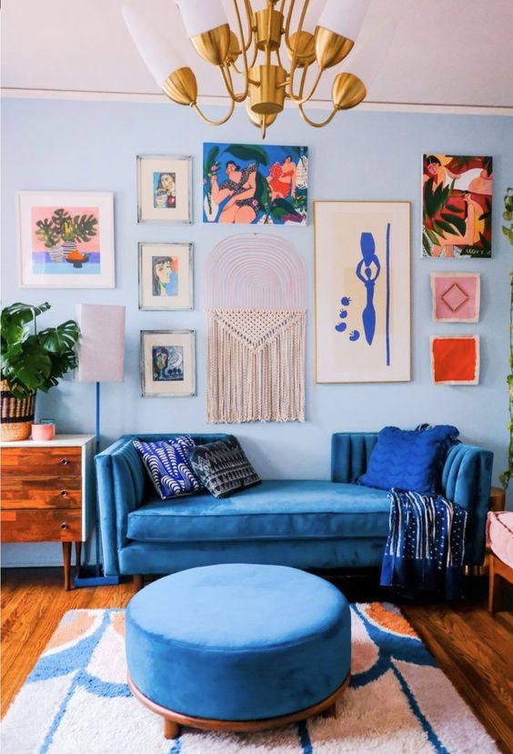 a bold maximalist living room with light blue walls, a pretty gallery wall, a blue sofa and an ottoman, some mid-century modern furniture