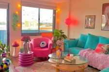 a bright and colorful maximalist living room with a turquoise sofa, a pink chair, a glass table, lights, plants and a bold prnted rug