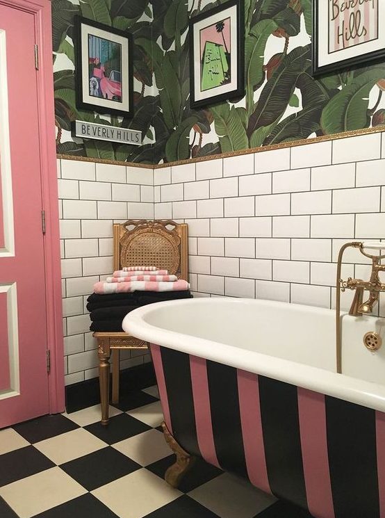 a bright bathroom with white subway tiles, tropical leaf wallpaper, a striped bathtub, a pink door and a checked floor plus pop artworks