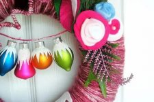 a bright pink Christmas wreath with white fabric styled as a donut, with colorful fabric blooms, colorful Christmas ornaments and evergreens is amazing