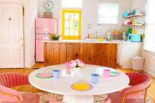 a candy-colored maximalist kitchen with a neon yellow window, a pink fridge, pink velvet chairs and pastel tableware