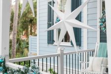a coastal Christmas porch with an evergreen garland with ornaments, white stars hanging over the space, white rockers with mint and aqua blankets