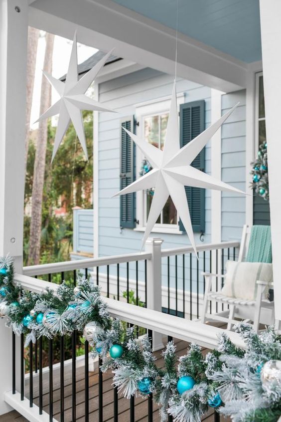 a coastal Christmas porch with an evergreen garland with ornaments, white stars hanging over the space, white rockers with mint and aqua blankets
