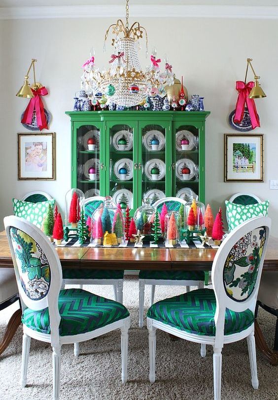 a colorful Christmas dining room with a green buffet with colorful ornaments in mugs, colorful bottle brush Christmas trees on the table and pink bows