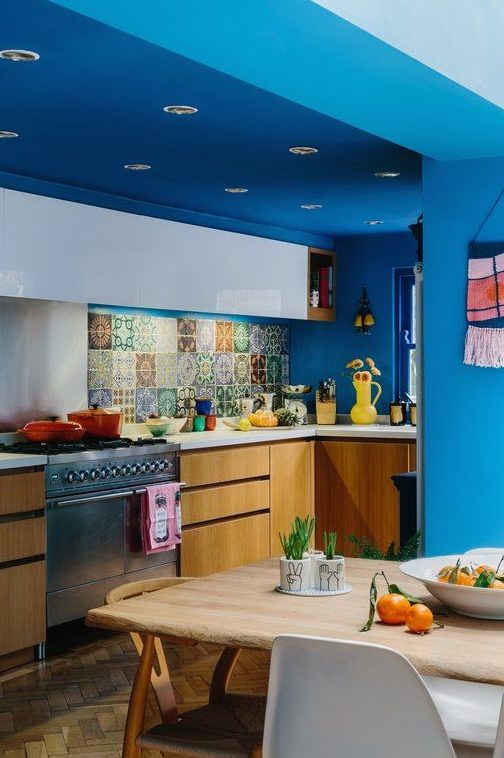 a colorful maximalist kitchen with bold blue walls and a ceiling, white and stained cabinets, white countertops and a colorful backsplash