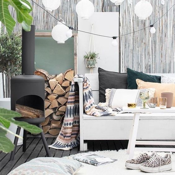 a cozy modern winter terrace with a white wooden bench with mismatching pillows and a folksy blanket, a hearth, firewood, some rugs and paper lamps