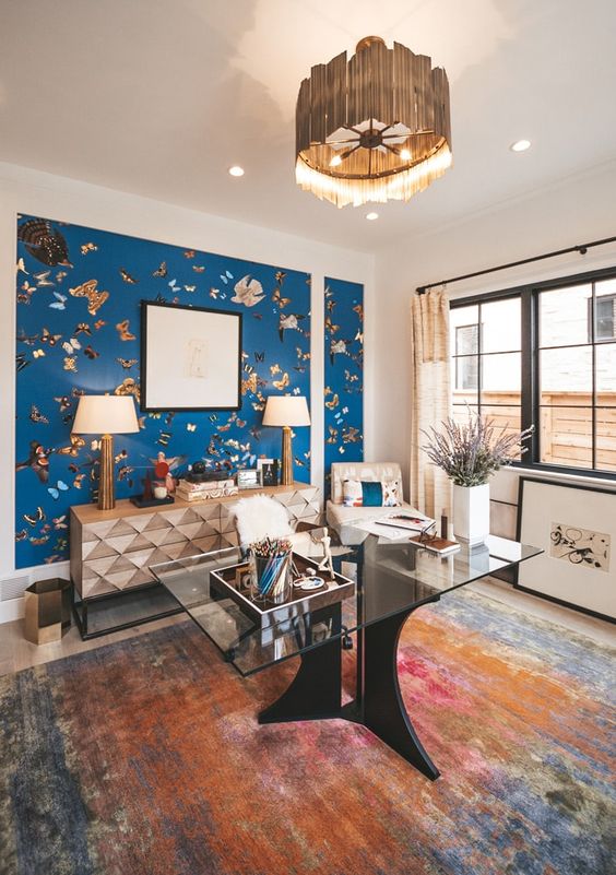 a creative maximalist home office with a blue printed accent wall, a faceted credenza, a clear glass desk, a chic metallic pendant lamp (designed by MARGARITA BRAVO)