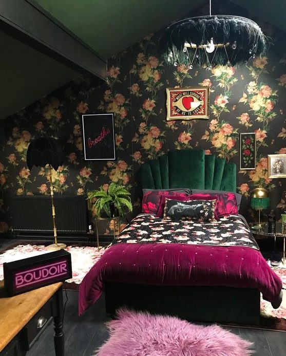a dark maximalist bedroom with a dark floral accent wall, a dark green velvet bed, a feather chandelier, bold floral and purple bedding and a neon light
