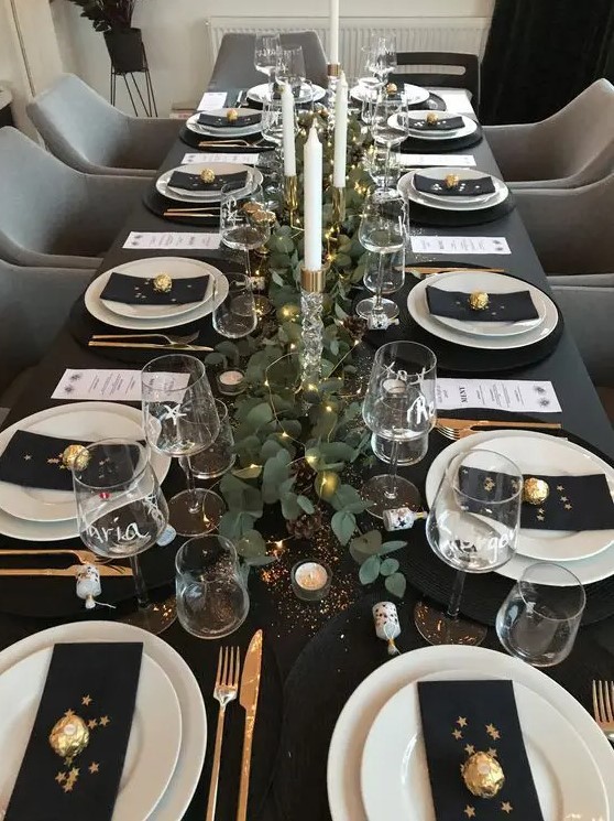 a dreamy tablescape in black, with black linens, white porcelain, a greenery runner with LED lights, tall and thin candles