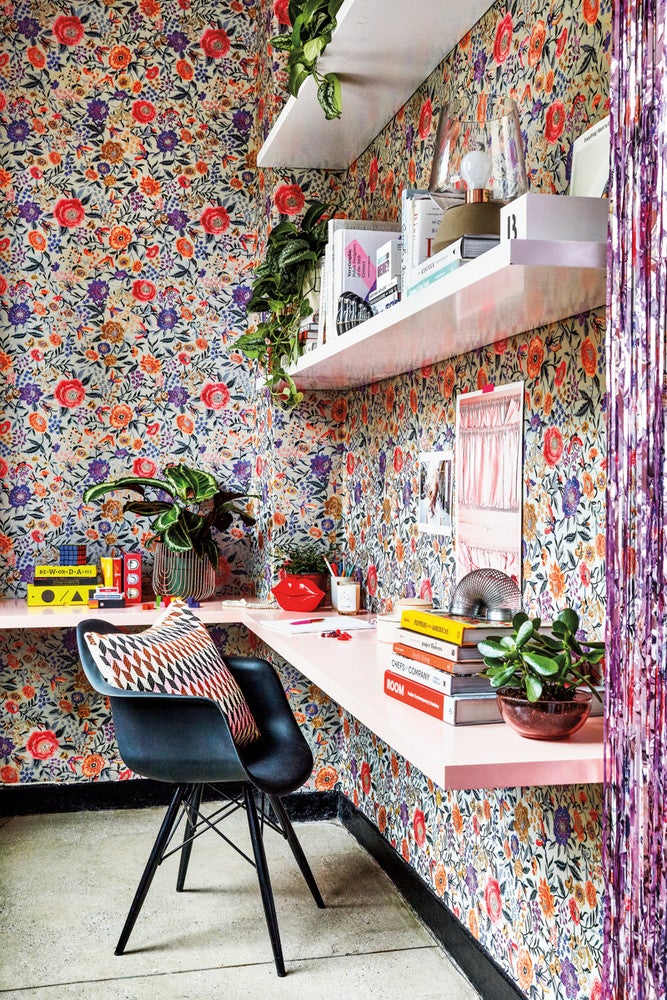 a jaw-dropping home office nook covered with bright floral wallpaper, white floating shelves and a pink floating desk, lots of books and potted plants