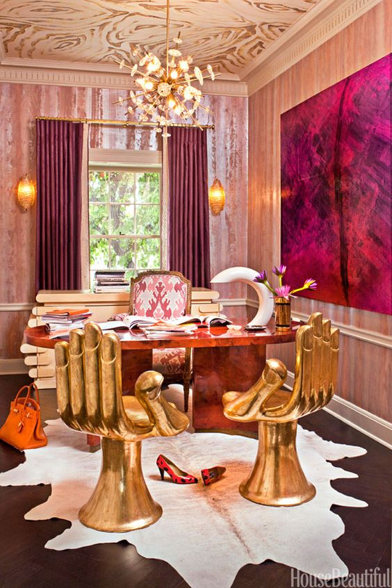 a jaw dropping maximalist home office wiht a patterned ceiling, pink wallpaper, a stone desk, gold hand shaped chairs, a statement artwork and curtains that echo with it