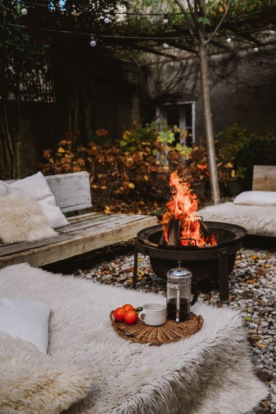 a lovely winter patio with pebbles on the ground, a metal fire bowl, wooden benches covered with faux fur and faux fur pillows