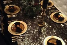 a luxurious glam Christmas tablescape with a shiny tablecloth, gold chargers and cutlery, black napkins and candles and a fir and pinecone centerpiece