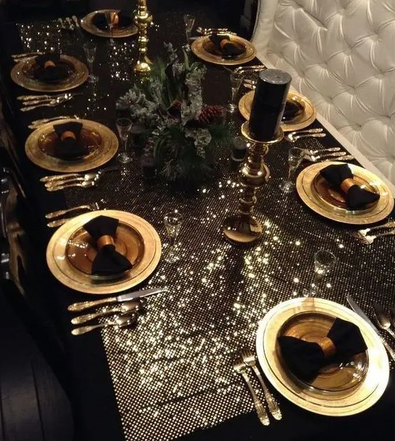 a luxurious glam Christmas tablescape with a shiny tablecloth, gold chargers and cutlery, black napkins and candles and a fir and pinecone centerpiece