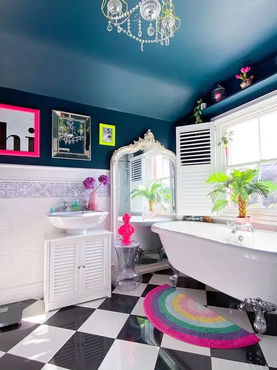a maximalist bathroom with a light blue ceiling, navy walls, white furniture and a clawfoot tub, mirrors, artworks and a bold rug
