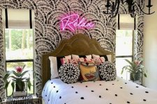 a maximalist bedroom with a printed accent wall, a green bed, bright bedding and a rug, a neon light and a black chandelier