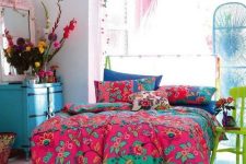 a maximalist bedroom with pink walls, a bed, a blue chest of drawers, a green chair, colorful bedding and bold blooms plus a lovely pendant lamp