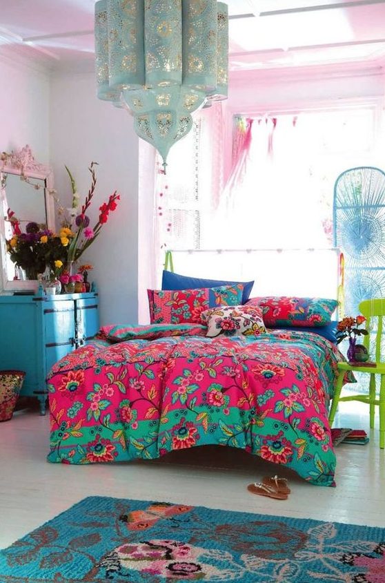 a maximalist bedroom with pink walls, a bed, a blue chest of drawers, a green chair, colorful bedding and bold blooms plus a lovely pendant lamp