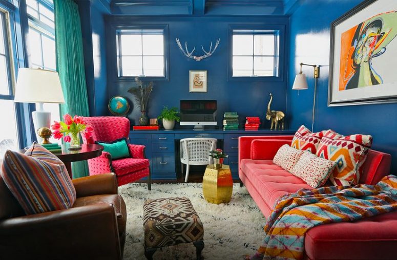 a maximalist home office with blue walls and a matching larrge desk, a coral daybed, mismatching chairs, green curtains and crazy touches of pattern