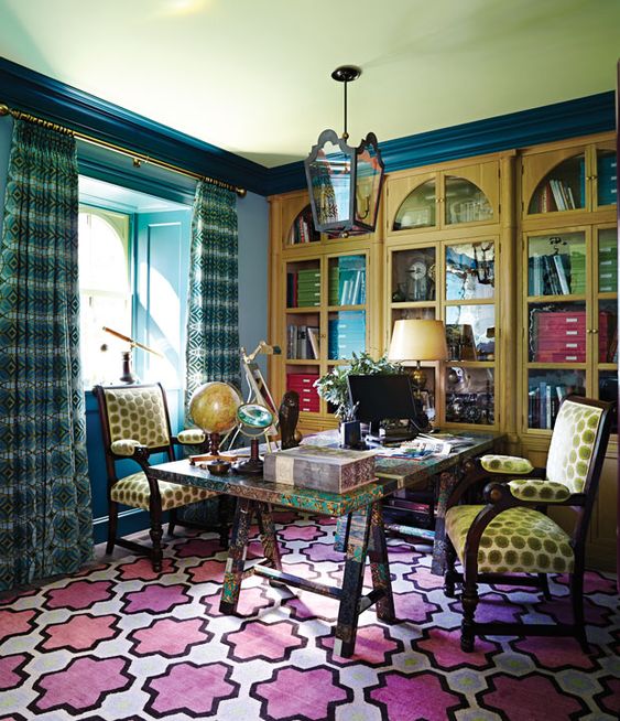 a maximalist home office with blue walls, yellow built-in bookcases, a colorful trestle desk and a colorful rug, bold mustard chairs