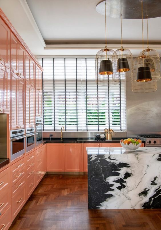 a maximalist kitchen in slamon pink, with a black marble slab kitchen island, black pendant lamps in cages and gold touches