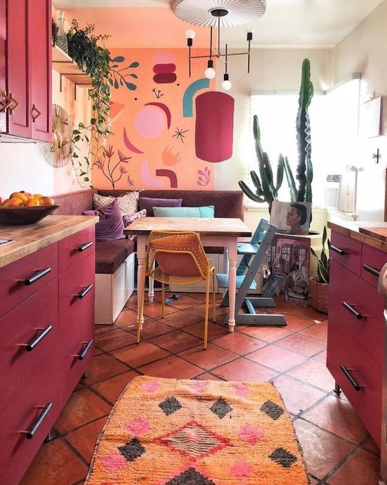 a maximalist kitchen with an orange wall, fuchsia cabinets, a mid century modern chandelier and bold textiles
