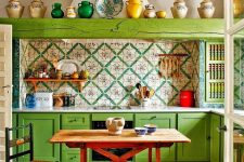 a maximalist kitchen with bold green cabinets and a wooden beam, bold printed tiles and geometric ones on the floor, a stained table and a woven chair