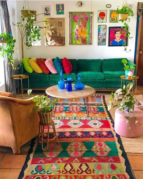 a maximalist living room with an emerald sofa, a bright printed rug, a colorful gallery wall, a rust colored chair, potted greenery and some blue accessories
