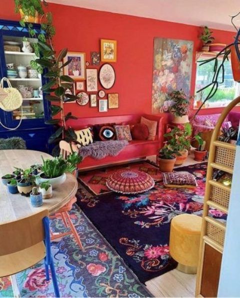 a maximalist living room with red walls and a matching sofa, layered floral print rugs, a navy buffet, lots of potted greenert and some artworks