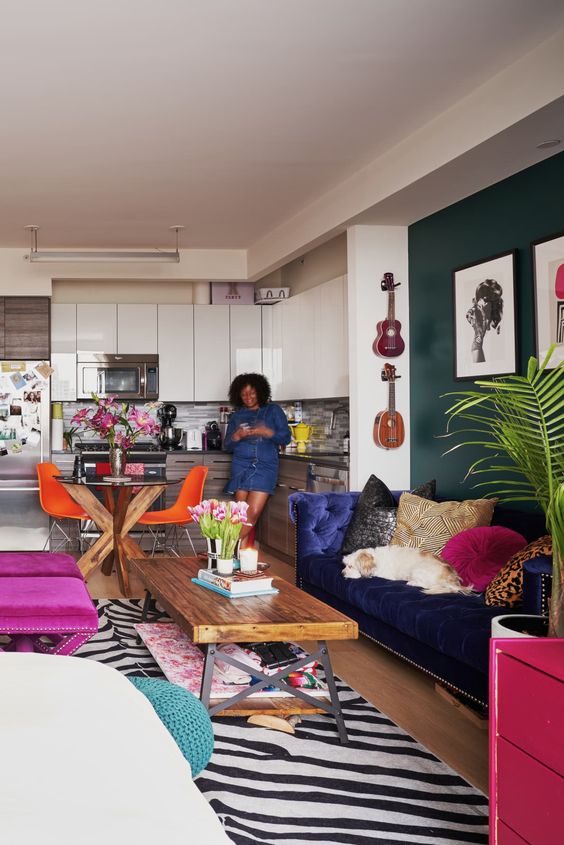 a maximalist open layout with a dark green accent wall, a navy sofa, colorful printed pillows, hot pink stools and a printed rug