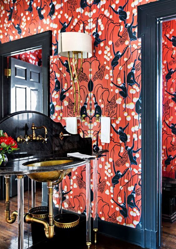 a mind blowing maximalist bathroom with red printed wallpaper, a black, gold and clear acryl vanity, a gold sink, some lamps and a mirror wall