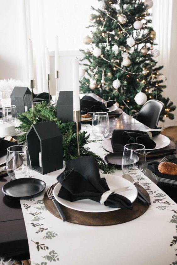 a modern black and white Christmas tablescape with a printed runner, black napkins, black house shaped candleholders and evergreens and white candles