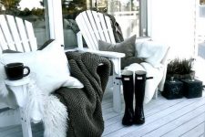 a monochromatic winter terrace with a white deck, white planked chairs, grey, black and white pillows and blankets, rubber boots and some greenery