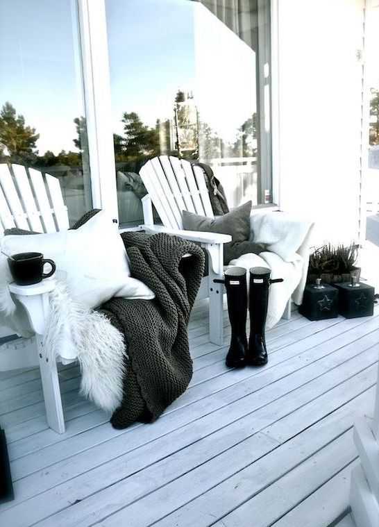a monochromatic winter terrace with a white deck, white planked chairs, grey, black and white pillows and blankets, rubber boots and some greenery