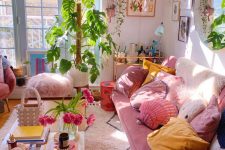 a pretty maximalist living room with blush walls, blue framed doors, a pink sofa, colorful pillows, pink seating furniture and greenery plus a gallery wall