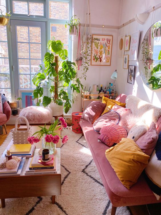 a pretty maximalist living room with blush walls, blue framed doors, a pink sofa, colorful pillows, pink seating furniture and greenery plus a gallery wall