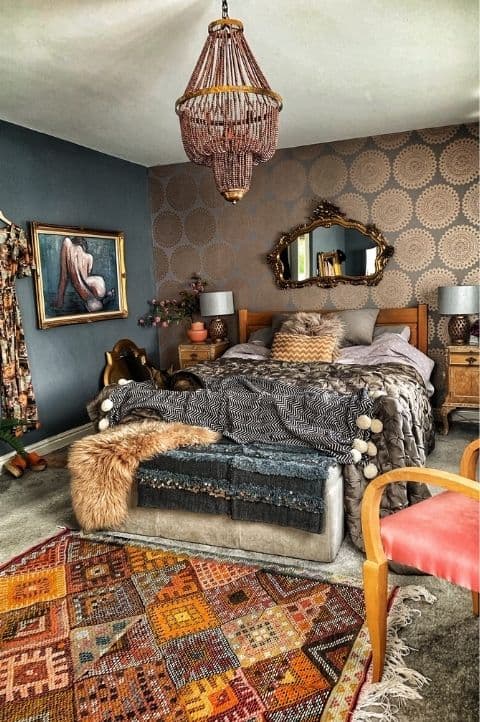 a refined dark maximalist bedroom with black walls and a metallic printed one, a wooden bed and layered bedding, a soft bench with faux fur and a beaded chandelier