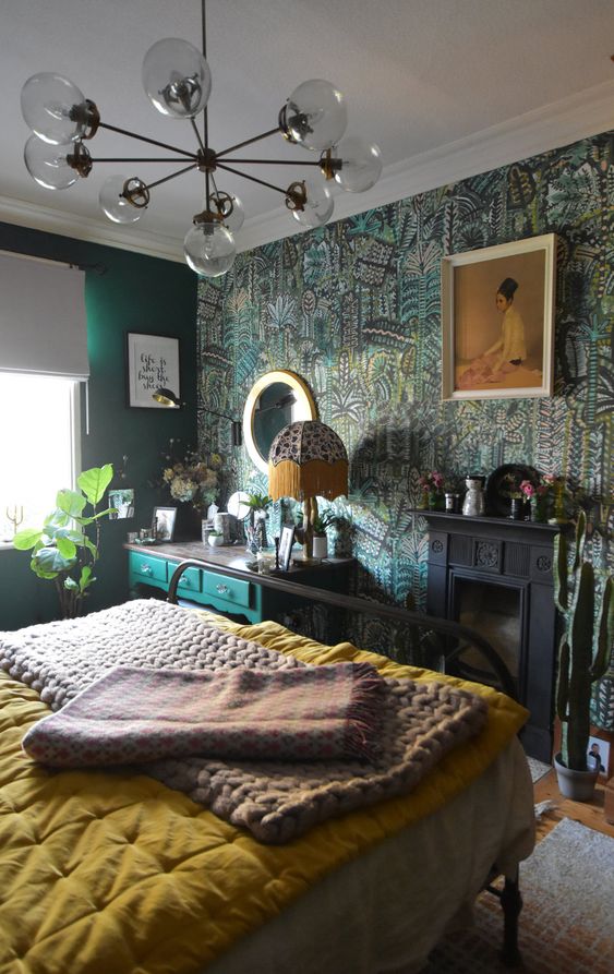 a refined green maximalist bedroom with a printed wallpaper accent wall, a black faux fireplace and a black forged bed, a green makeup table and some plants