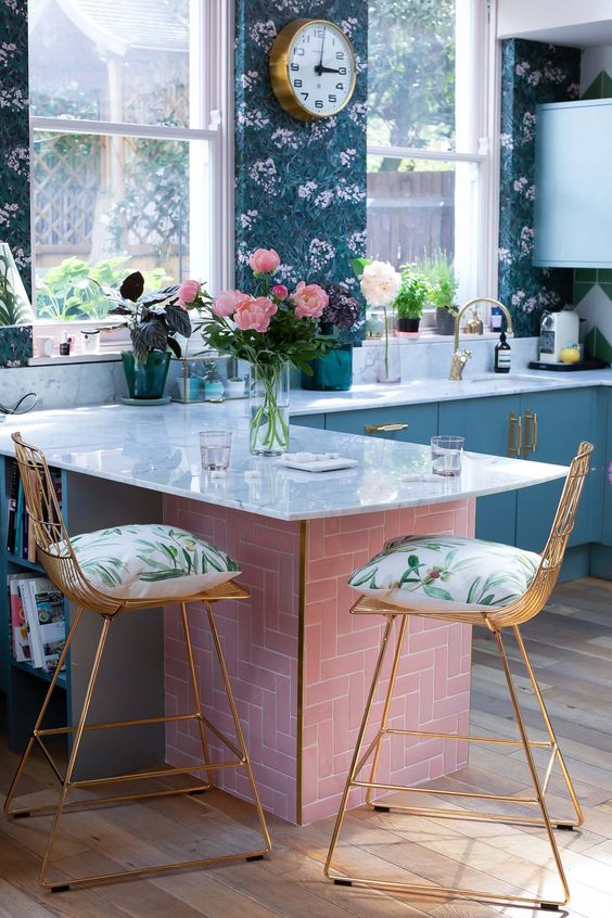a refined maximalist kitchen with moody floral wallpaper walls, pale blue cabinets, a pink kitchen island with a white stone countertop and tall stools with printed cushions