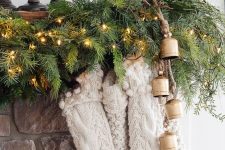a rustic Christmas mantel with an evergreen and lights garland, white stockings, vintage bells is a very cozy idea