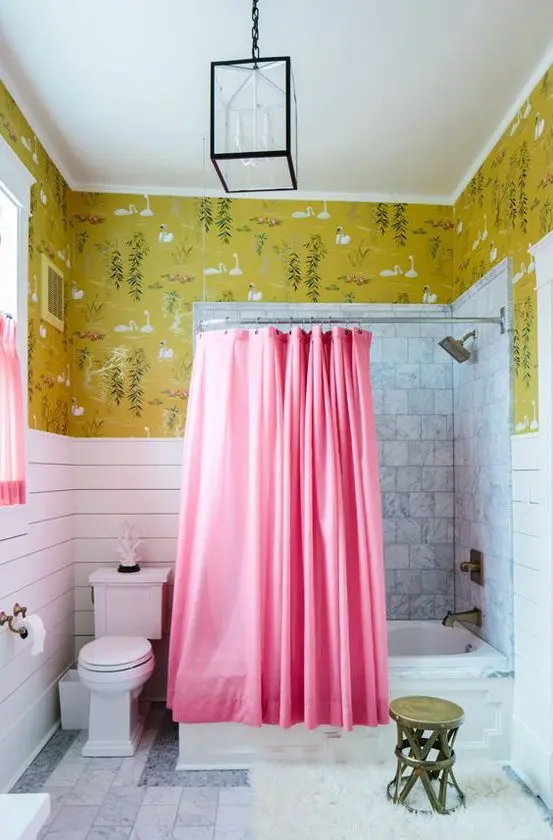 a stylish bathroom with bold mustard wallpaper, white paneling and marble tiles, pink curtains and a brass stool and fixtures just wows