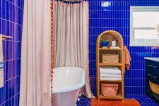 a super vibrant maximalist bathroom clad with electric blue tiles, a vintage tub with a large curtain, an orange rug, a rattan shelf and a black vanity